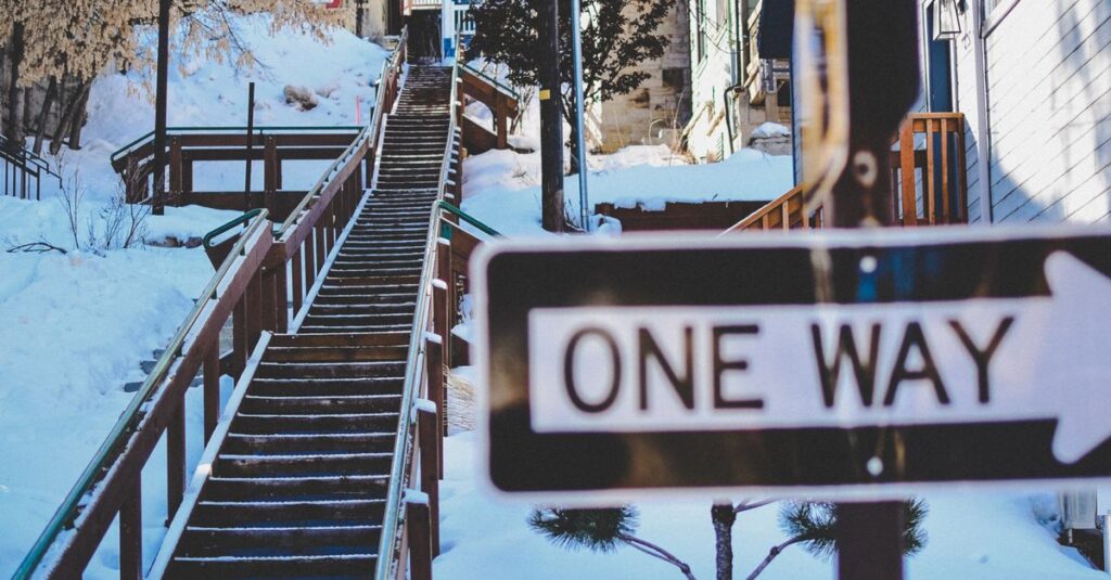 Snow-covered staircase leading uphill with a visible one-way sign in a winter cityscape.